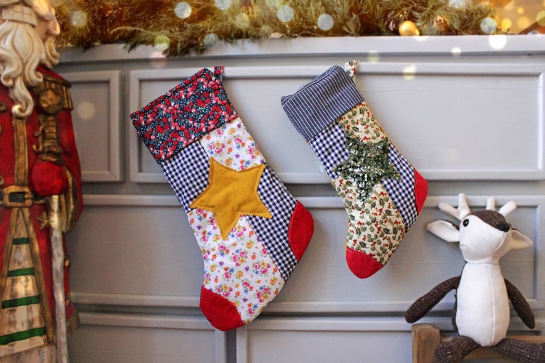 Pode incluir: Duas meias de Natal feitas de tecido patchwork com designs diferentes. Uma meia tem uma estrela amarela, a outra tem uma estrela verde com lantejoulas. Ambas as meias t&ecirc;m uma borda vermelha.