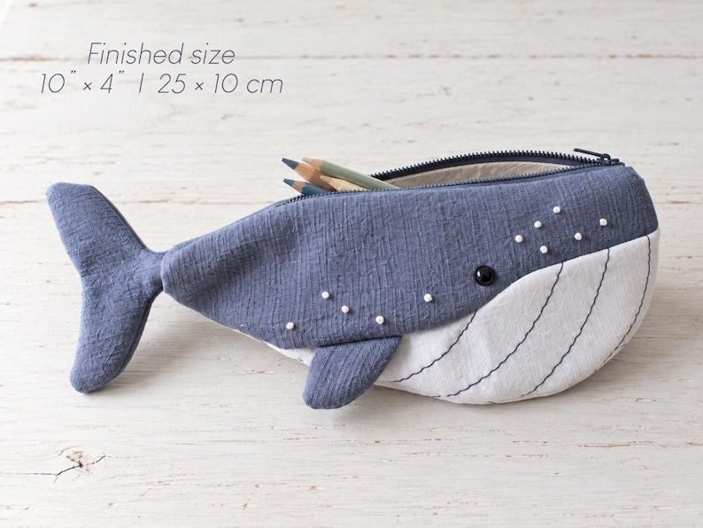 Pu&ograve; includere: Un astuccio a forma di balena in tessuto blu e bianco con chiusura a zip. L'astuccio presenta piccole perline bianche decorative e linee nere cucite sulla parte inferiore bianca. La dimensione finita &egrave; di 25 cm per 10 cm.