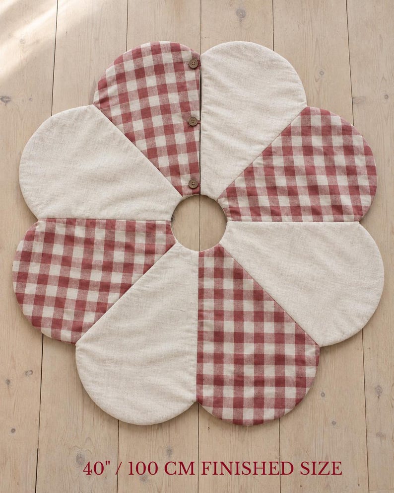 K&ouml;nnte beinhalten: Ein blumenf&ouml;rmiger Baumteppich mit einer fertigen Gr&ouml;&szlig;e von 100 cm, bestehend aus abwechselnden Bl&uuml;tenbl&auml;ttern aus beigem Stoff und rot-wei&szlig;em Vichy-Karo-Stoff, befestigt mit drei kleinen braunen Kn&ouml;pfen an einer Naht.
