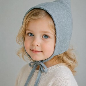 May include: A light blue pixie hat with a pointed top and chin ties. The text reads: PIXIE HAT PDF SEWING PATTERN 11 SIZES NEWBORN TO ADULT XL. The hat features a textured fabric construction that covers the head and secures with a bow under the chin.