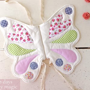 May include: Butterfly wings made of white fabric with quilted sections featuring strawberry, green gingham, pink striped, blue floral, and multi-colored floral patterns. The wings include cream-colored fabric ties and the text: For dress-up days and everyday magic.