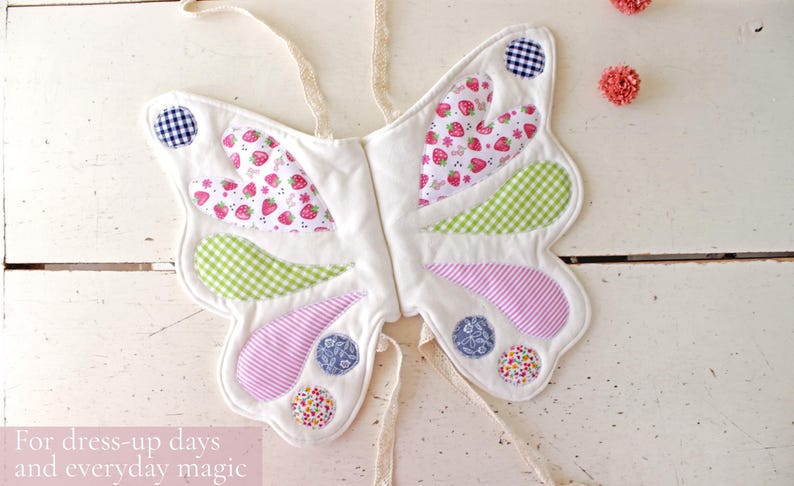 May include: Butterfly wings made of white fabric with quilted sections featuring strawberry, green gingham, pink striped, blue floral, and multi-colored floral patterns. The wings include cream-colored fabric ties and the text: For dress-up days and everyday magic.