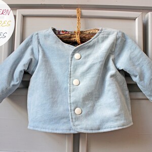 May include: A light blue long-sleeve jacket with three white buttons down the front. A circular white graphic in the top left corner contains the text PDF PATTERN +40 PICTURES +20 PAGES. The jacket is hung on a rustic wooden hanger.