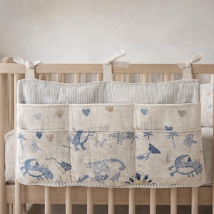 May include: A beige fabric nursery organizer with three pockets, adorned with blue dinosaur illustrations and hearts. The organizer hangs from a wooden crib, offering a charming storage solution for baby essentials.