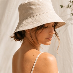 May include: A solid light beige bucket hat with a flat crown and a downward-sloping brim. The hat features a visible woven texture throughout the fabric, constructed with vertical seams along the sides and horizontal stitching around the brim.