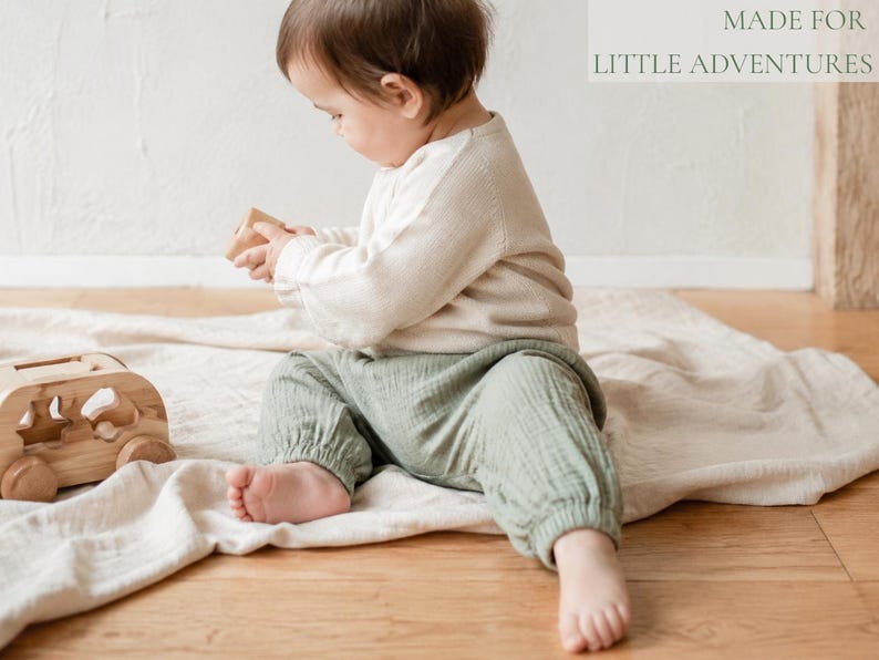 May include: A child wearing a long-sleeved cream-colored top and sage green muslin pants, sitting on a light-colored fabric. Beside the child is a wooden toy vehicle. The text "MADE FOR LITTLE ADVENTURES" is visible in the upper right corner.