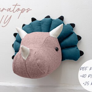 May include: A DIY Triceratops head wall decor pattern with 40 pictures and 25 pages. The pattern includes a pink felt head with white horns and a teal felt frill.