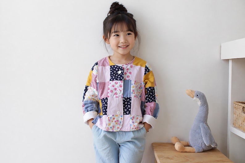 May include: A long-sleeved jacket featuring a patchwork design with various fabric squares in shades of pink, yellow, blue, and white, including floral and striped patterns. The jacket has a round neckline, a front button closure, and white cuffs.
