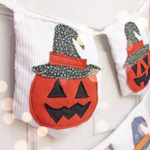May include: A white fabric banner with three orange and black felt pumpkin decorations. Each pumpkin has a black felt hat with a floral pattern.