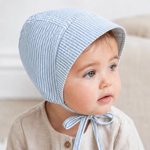 Può includere: Una cuffietta per neonati con un motivo a righe verticali sottili color azzurro e bianco. La cuffietta è realizzata in tessuto seersucker testurizzato e include lacci in tessuto abbinati che si allacciano sotto il mento per fissare il copricapo.