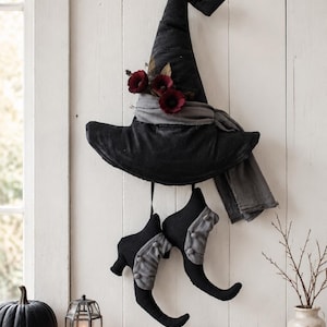 May include: A black fabric witch hat with a pointed top and a wide brim, adorned with a grey fabric band and three dark red flowers. Two black fabric boots with grey cuffs and small grey buttons hang from the hat by thin black straps.