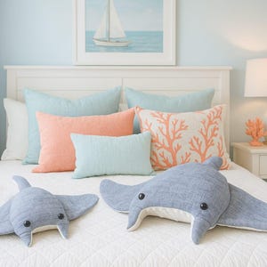 May include: A bedroom scene with a white bed, featuring various throw pillows in shades of blue, coral, and white. Two gray stingray-shaped plush toys are on the bed. A framed sailboat art piece hangs above the bed.