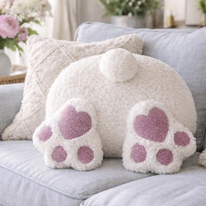 May include: A plush, white bunny-shaped pillow with a fluffy tail and two paws with pink heart-shaped pads. The pillow is on a light blue sofa, with other decorative pillows and flowers in the background.