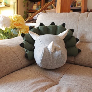 May include: A plush gray and green dinosaur head pillow with a white horn. The pillow is sitting on a beige couch.