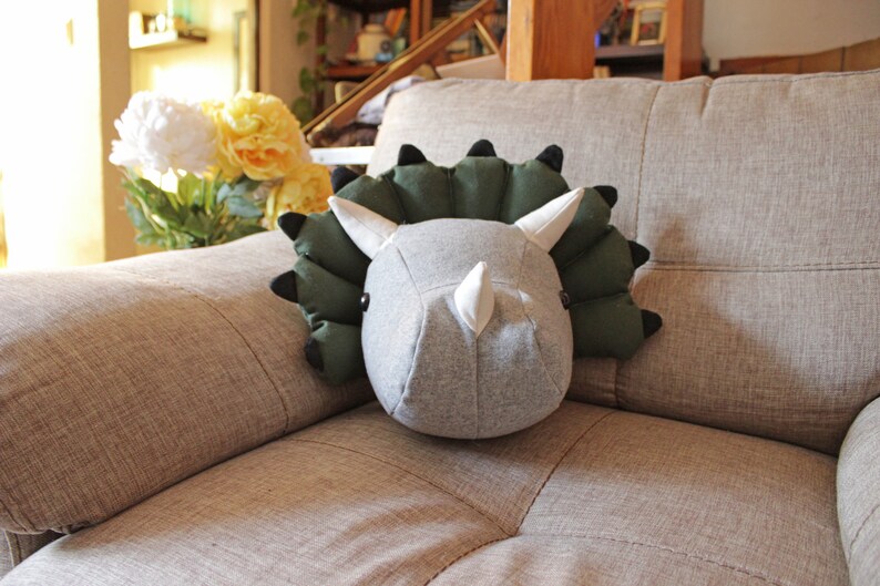 May include: A plush gray and green dinosaur head pillow with a white horn. The pillow is sitting on a beige couch.