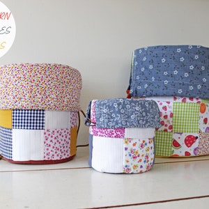 May include: Three fabric storage baskets in different sizes. The largest basket is blue with white polka dots, the middle basket is white with a patchwork design of pink, blue, and white floral patterns, and the smallest basket is green with a patchwork design of red, white, and green fruit patterns. The baskets are on a white wooden surface.