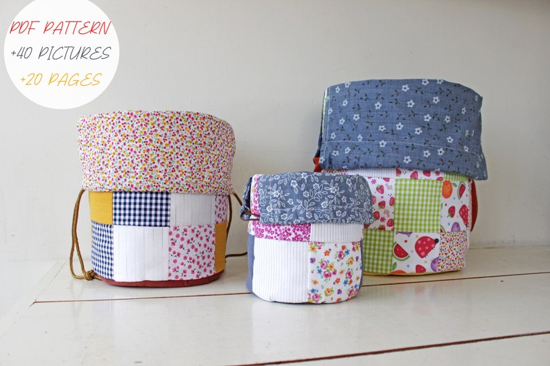 May include: Three fabric storage baskets in different sizes. The largest basket is blue with white polka dots, the middle basket is white with a patchwork design of pink, blue, and white floral patterns, and the smallest basket is green with a patchwork design of red, white, and green fruit patterns. The baskets are on a white wooden surface.