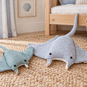 May include: Two plush stingray toys featuring soft fabric bodies with black button eyes. One stingray is solid light blue, while the larger one features a blue and white vertical striped pattern. Both have long tails and wide fins.