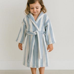 Può includere: Un accappatoio a righe blu e bianche per bambini, con cappuccio e cintura annodata. L'accappatoio è realizzato in un materiale morbido e felpato. Il bambino è in piedi a piedi nudi.