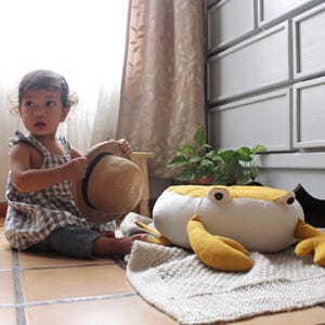 May include: A child in a checkered dress holds a straw hat. A large, plush crab toy with yellow claws and a yellow top sits on a white rug. The background features a window, curtains, and a chest of drawers.