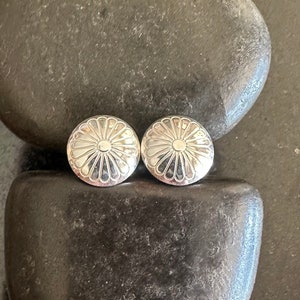 May include: A pair of silver earrings with a flower design. The earrings are round and have a raised center with a circle in the middle. The earrings are on a dark gray background.