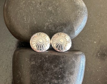 May include: A pair of silver earrings with a flower design. The earrings are round and have a raised center with a circle in the middle. The earrings are on a dark gray background.