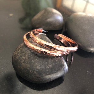 May include: Two copper-colored, hammered-texture hoop earrings. The earrings are open at the bottom and rest on a dark gray stone. The earrings have a shiny finish and a simple, elegant design. The background includes other dark gray stones.