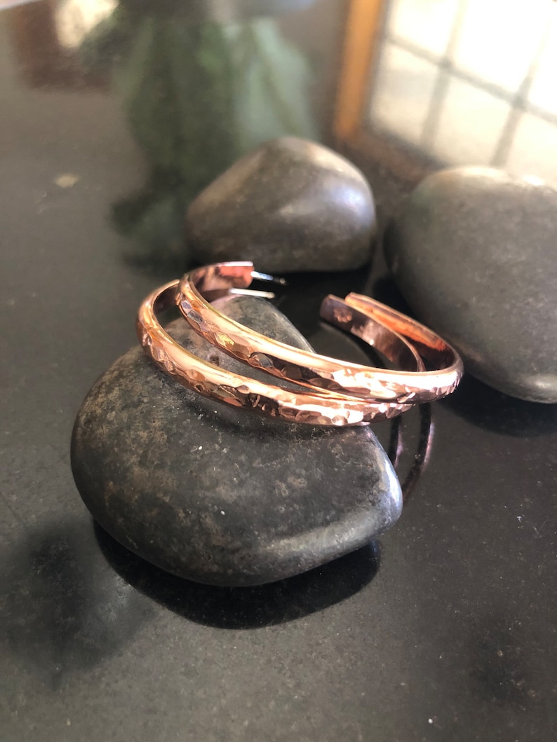 May include: Two copper-colored, hammered-texture hoop earrings. The earrings are open at the bottom and rest on a dark gray stone. The earrings have a shiny finish and a simple, elegant design. The background includes other dark gray stones.