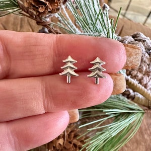 May include: A pair of silver-colored stud earrings shaped like Christmas trees. Each tree has a layered design with a small trunk. The earrings are displayed against a natural background with pine needles and a pine cone.