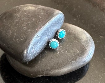 May include: Two turquoise stud earrings with a silver-coloured, scalloped bezel setting. The earrings are presented on a dark grey, textured stone surface. The bright blue-green stones contrast with the neutral background.
