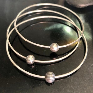 May include: Three silver bangle bracelets with different textured ball accents. The bracelets are stacked on top of each other.