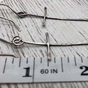 May include: A pair of silver-toned cross earrings. The earrings have a hammered texture and are approximately 2 inches long. The cross design is simple and elegant, with a hook closure.