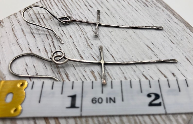 May include: A pair of silver-toned cross earrings. The earrings have a hammered texture and are approximately 2 inches long. The cross design is simple and elegant, with a hook closure.
