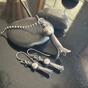 May include: A set of silver jewelry featuring a pendant necklace and matching earrings. The pendant is a stylized flower with a round bead at the top. The earrings are similar in design, with a smaller version of the flower pendant.