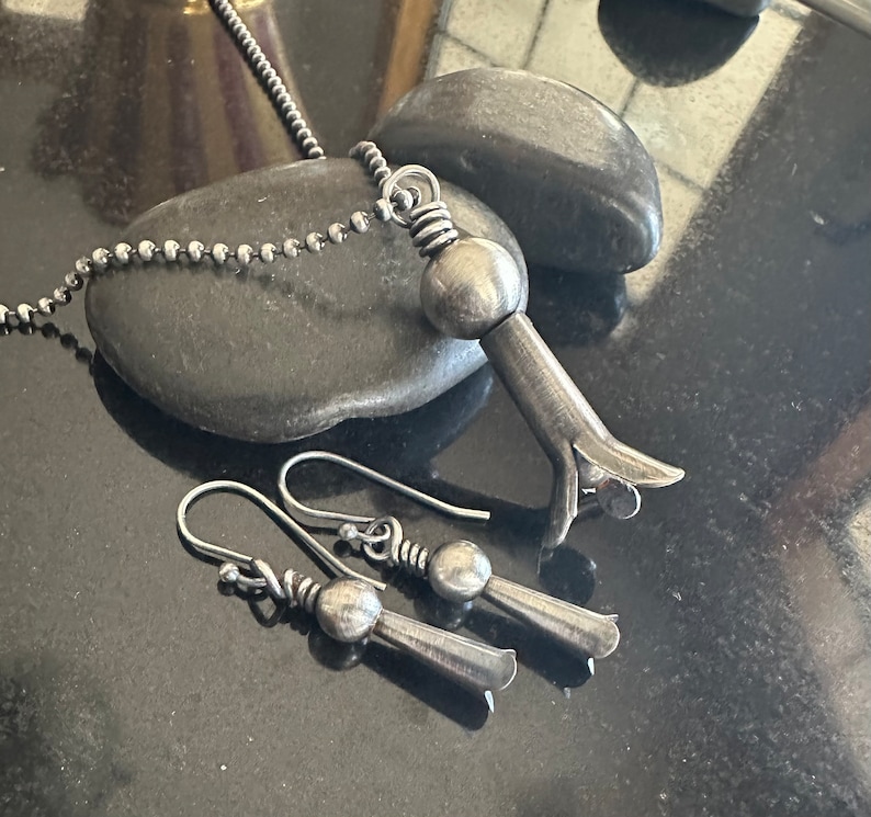 May include: A set of silver jewelry featuring a pendant necklace and matching earrings. The pendant is a stylized flower with a round bead at the top. The earrings are similar in design, with a smaller version of the flower pendant.