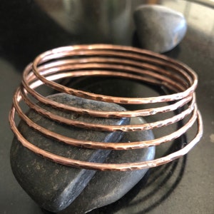May include: Set of six hammered copper bangles. The bangles are stacked on top of each other and have a warm, metallic sheen.