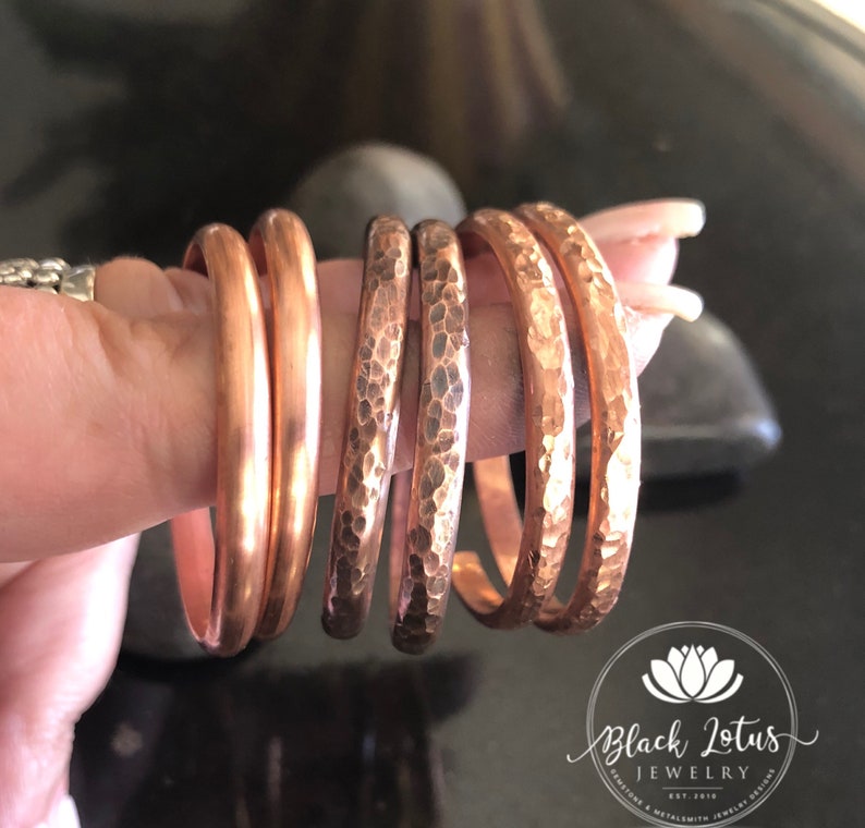 May include: A collection of copper bangles in varying widths and textures. Some are smooth, while others feature a hammered finish. The jewelry is displayed against a dark background, with the Black Lotus Jewelry logo visible.