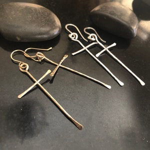 May include: Close-up of three pairs of cross-shaped earrings. Two pairs are silver, and one is gold. The earrings are displayed on a dark, reflective surface with two black stones in the background. The earrings have a hammered texture.