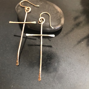 May include: A pair of gold-colored cross earrings. The earrings are handmade with a hammered texture and feature a simple cross design. The hooks are a looped design. The earrings are displayed on a dark gray stone.