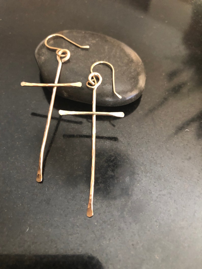 May include: A pair of gold-colored cross earrings. The earrings are handmade with a hammered texture and feature a simple cross design. The hooks are a looped design. The earrings are displayed on a dark gray stone.