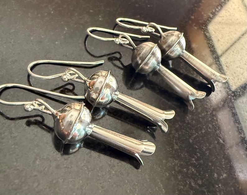 May include: Four pairs of silver earrings. Each earring has a rounded top with a textured surface and a long, slender stem ending in a flared, petal-like shape. The earrings have a hook-style closure.
