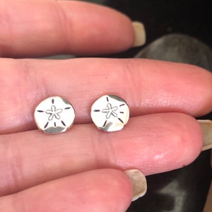 Könnte beinhalten: Ein Paar silberne Sanddollar-Ohrstecker. Die Ohrringe sind klein und rund, mit einem erhabenen Sanddollar-Design.