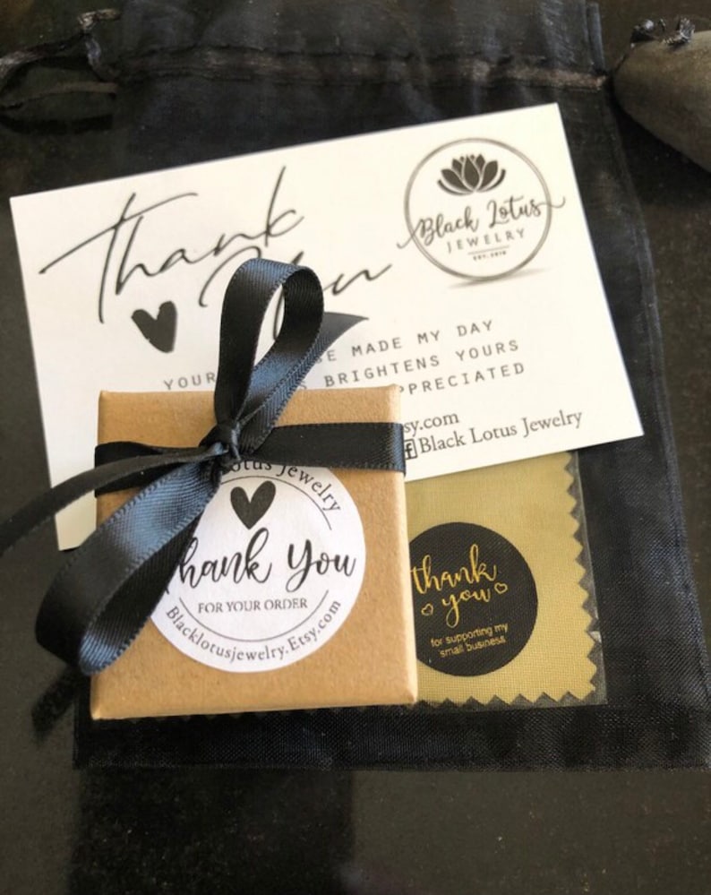 May include: A small, square, brown gift box tied with a black ribbon, alongside a white card with "thank you" and a black heart. The card also displays the text "Black Lotus Jewelry". A black pouch and a gold cleaning cloth are also visible.