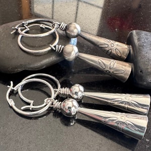 May include: Two pairs of silver hoop earrings with dangling charms. The charms are silver and have a textured, floral design.