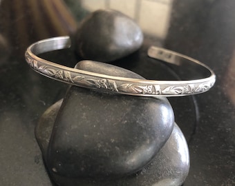 May include: A silver bangle bracelet with a delicate floral design. The bracelet is made of polished metal and has a simple, elegant style.