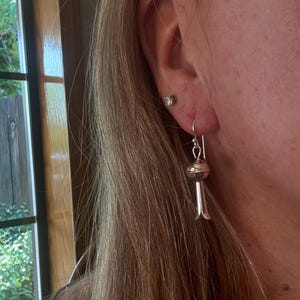 May include: Silver earrings with a distinctive design. Each earring has a small, round bead at the top, connected to a long, slender, bell-shaped dangle. The earrings are suspended from a silver hook. A small, clear stud earring is also visible.