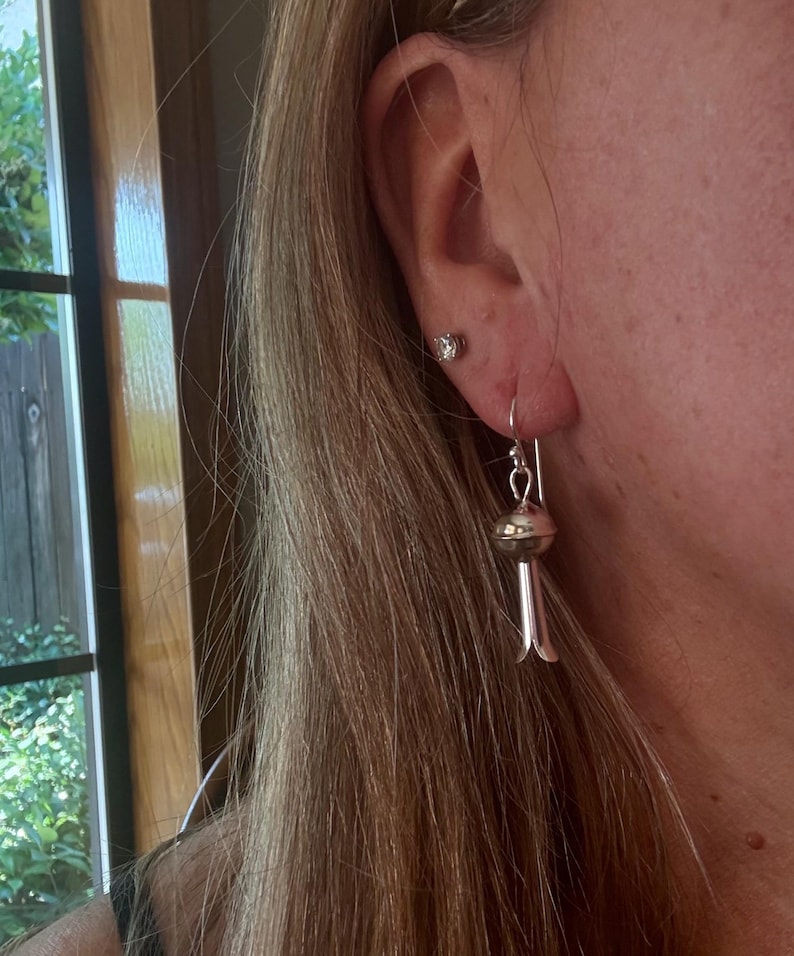 May include: Silver earrings with a distinctive design. Each earring has a small, round bead at the top, connected to a long, slender, bell-shaped dangle. The earrings are suspended from a silver hook. A small, clear stud earring is also visible.