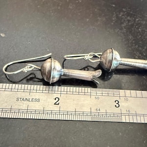 May include: A pair of silver earrings with a floral design. Each earring has a rounded, textured top and a slender stem with a flared end. The earrings are shown on a dark surface next to a ruler. The ruler is marked in inches.