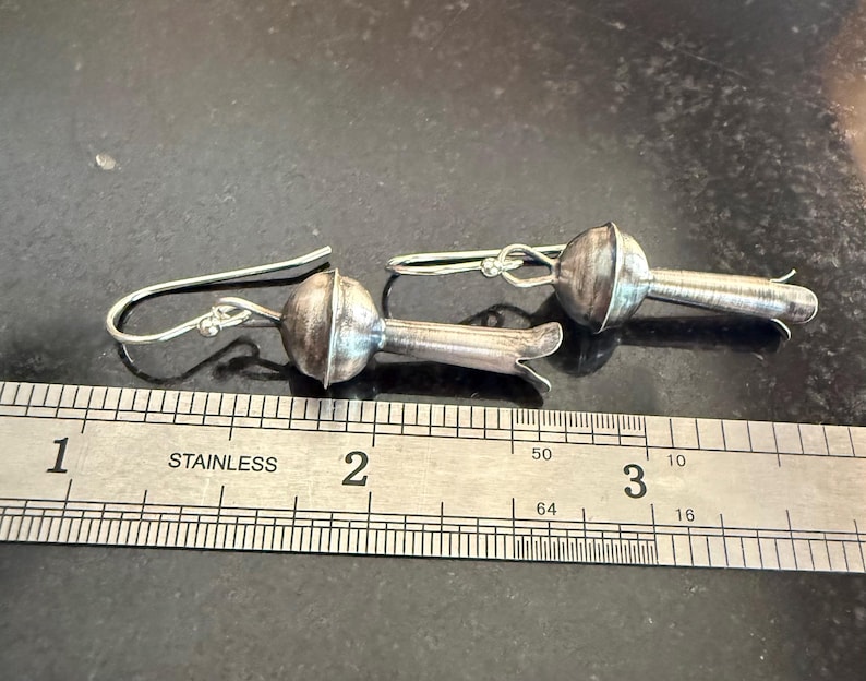 May include: A pair of silver earrings with a floral design. Each earring has a rounded, textured top and a slender stem with a flared end. The earrings are shown on a dark surface next to a ruler. The ruler is marked in inches.
