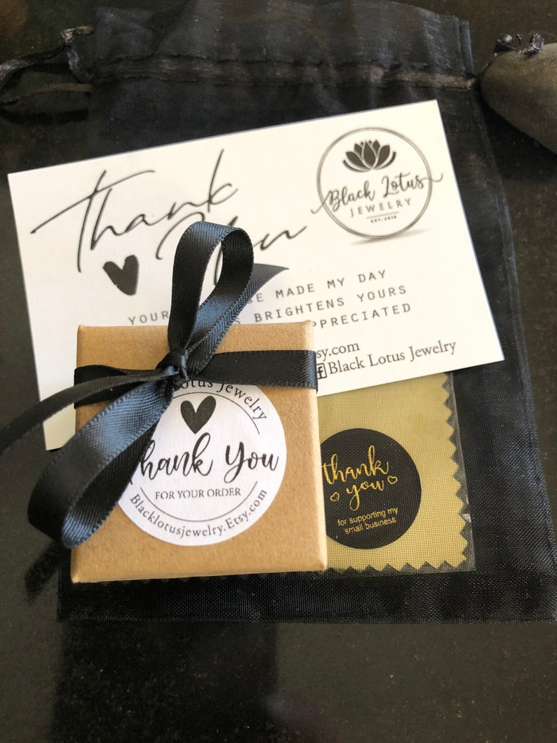 May include: A small, square, brown jewelry box tied with a black ribbon, with a "thank you" sticker. A white card with "Thank You" and "Black Lotus Jewelry" is visible, along with a black drawstring bag.